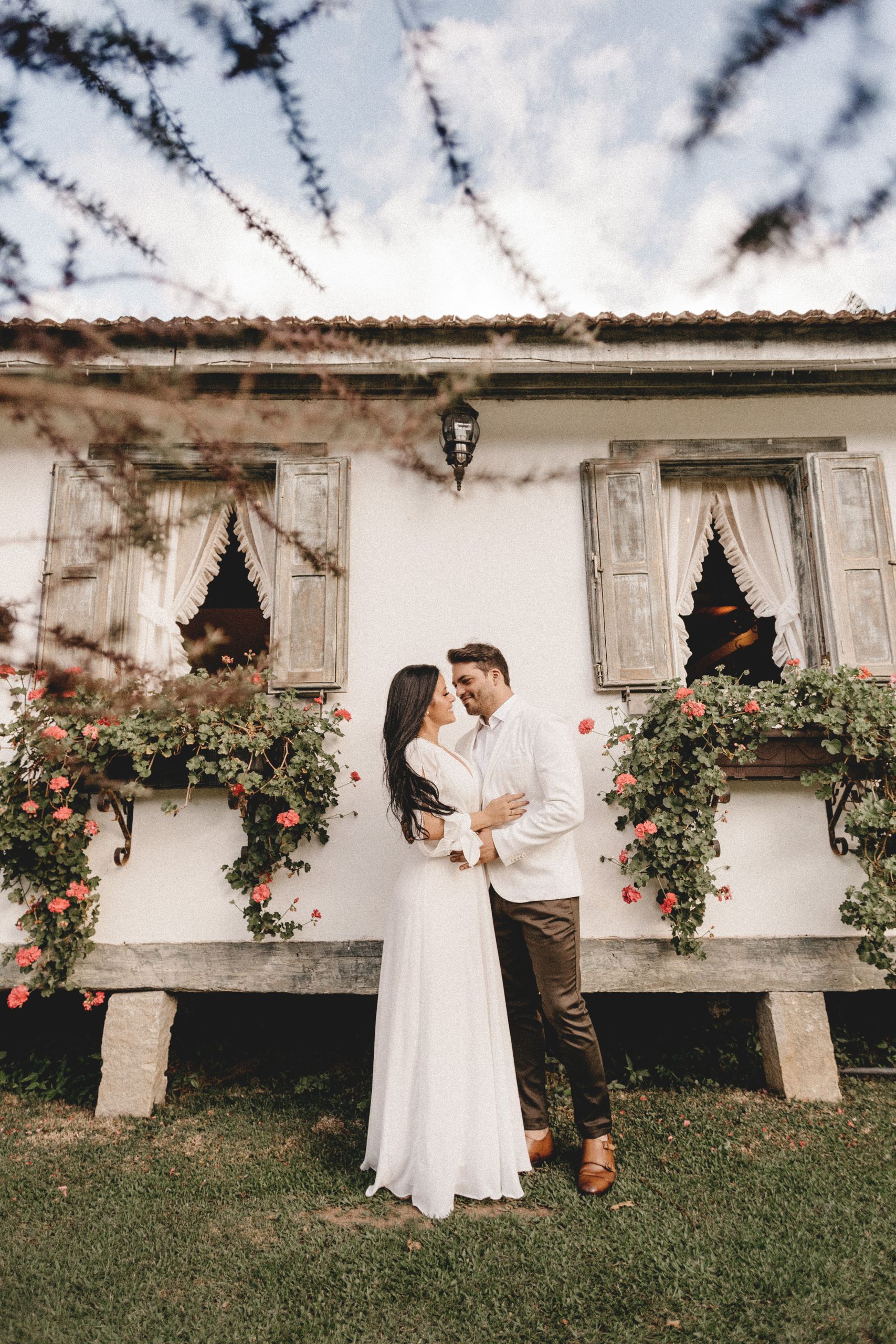 Low angle full body of smiling married couple wearing elegant clothes hugging gently against village house